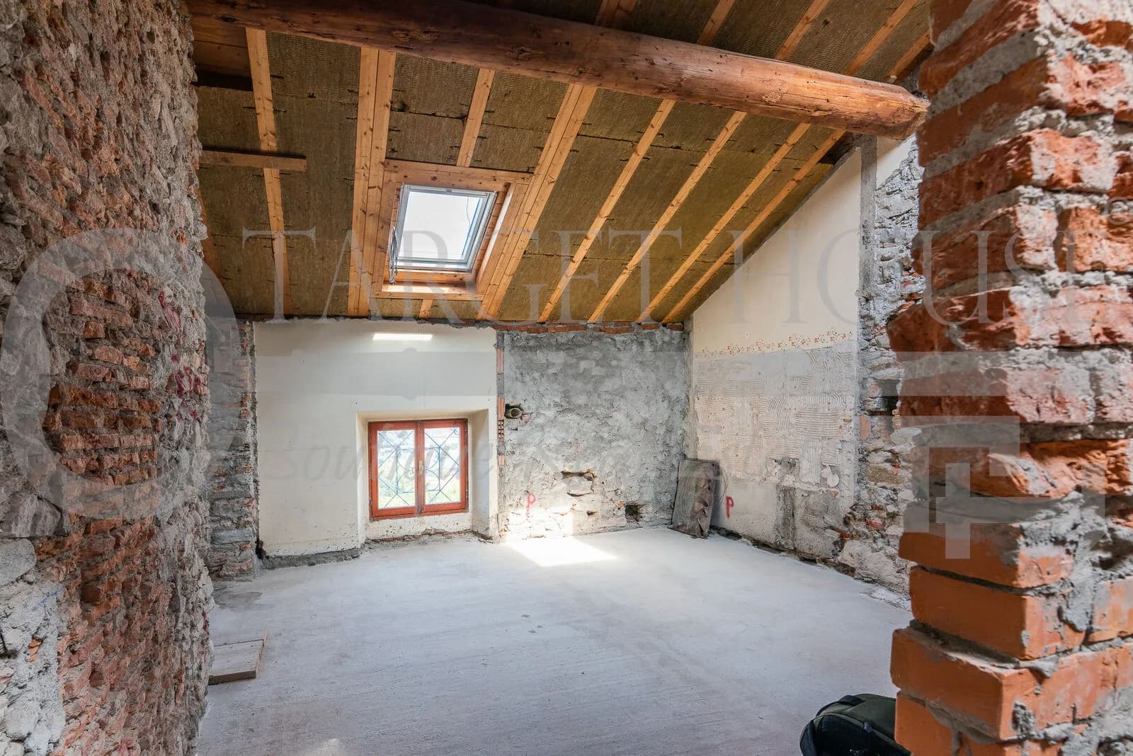 Attic room under renovation with exposed wooden beams, skylight, stone walls, and small arched window in historic villa near Como