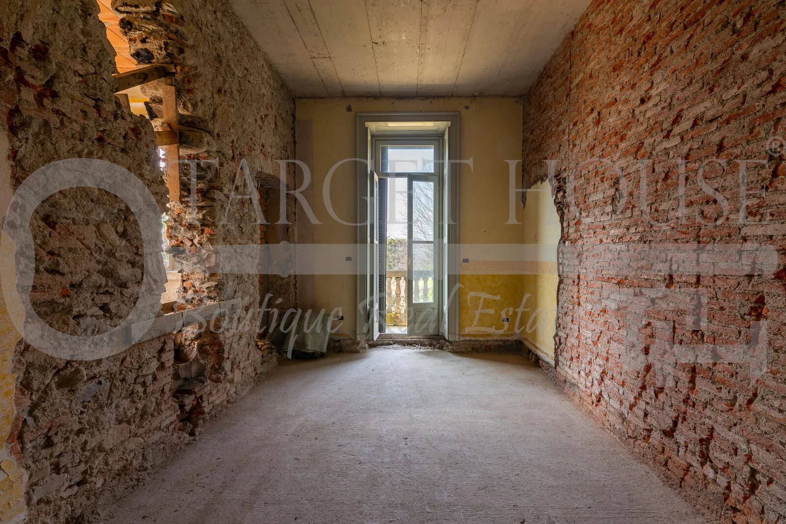 Stripped brick interior room with concrete floor and tall French door opening to balustrade balcony in historic villa near Como