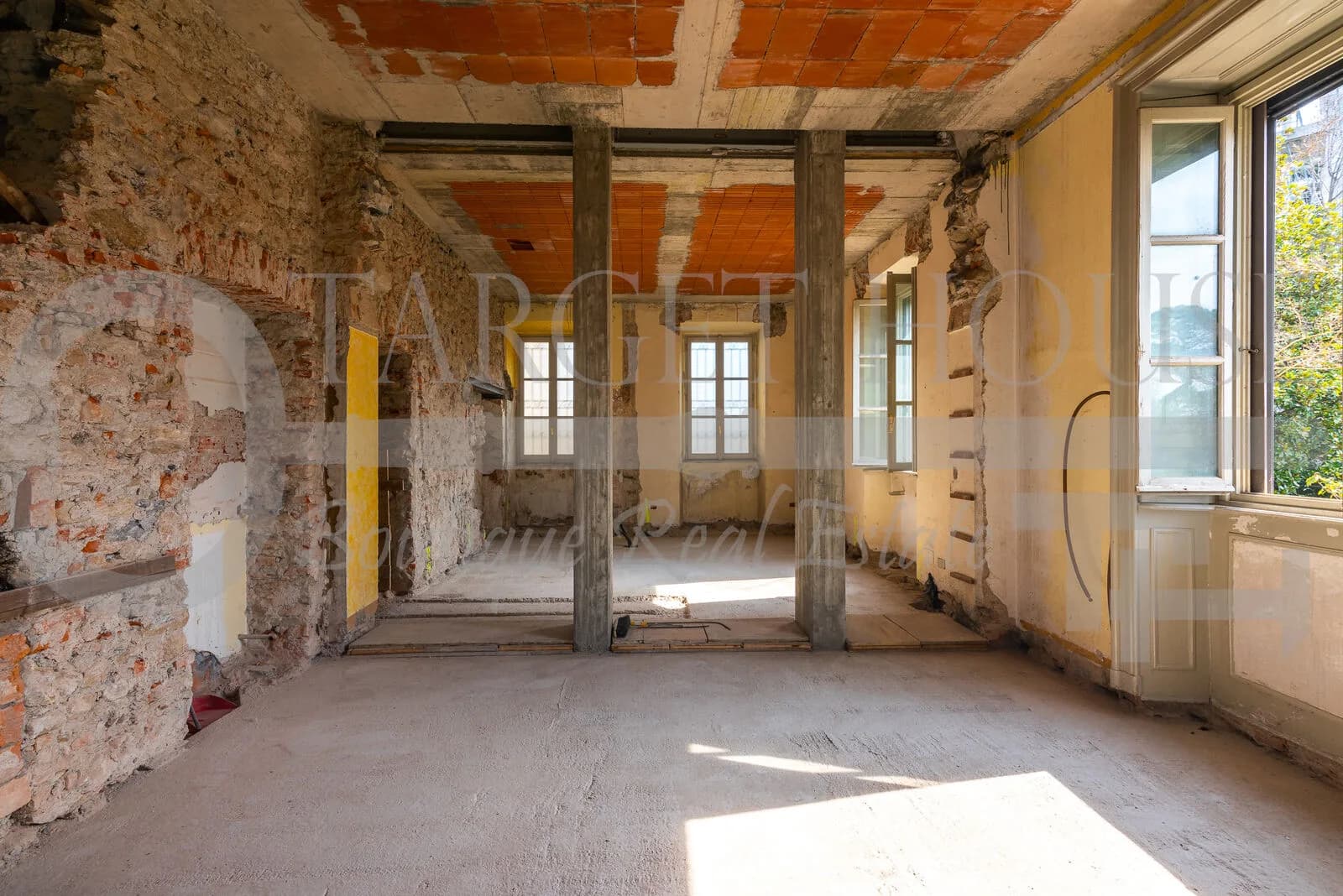 Interior of historic villa near Como mid-renovation, exposing brick walls, concrete columns, terracotta ceiling, and dusty bare floors