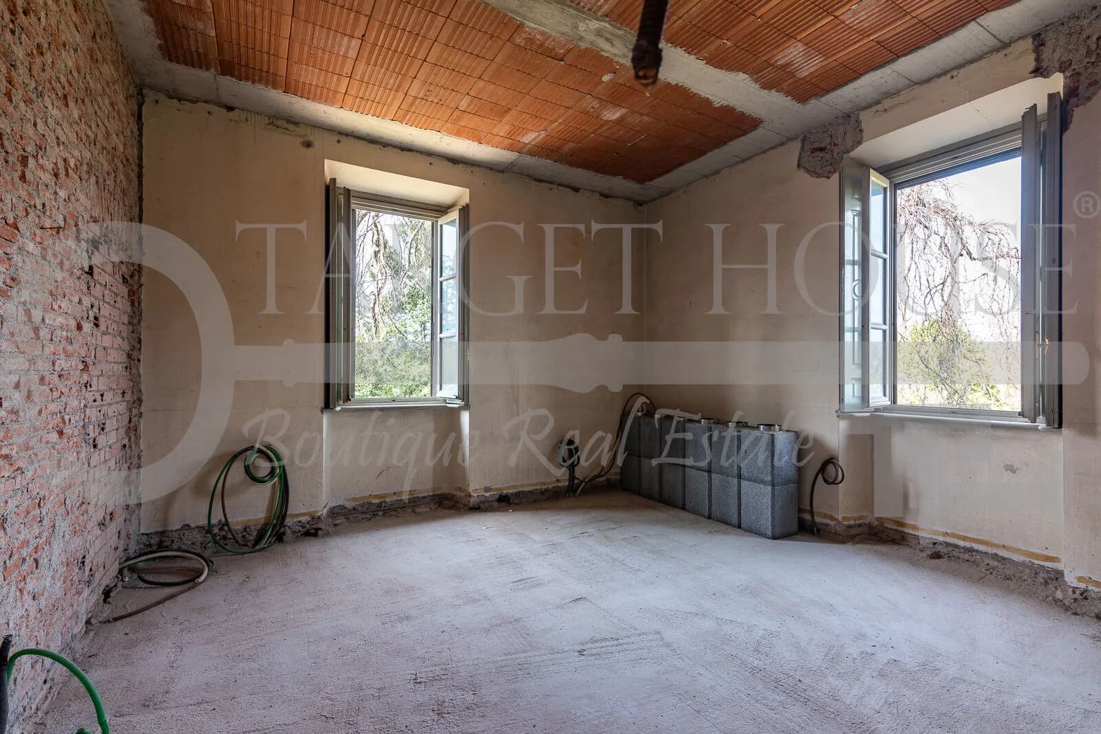 Unfinished interior room of historic villa near Como with exposed brick wall, terracotta ceiling, and dual garden-view windows