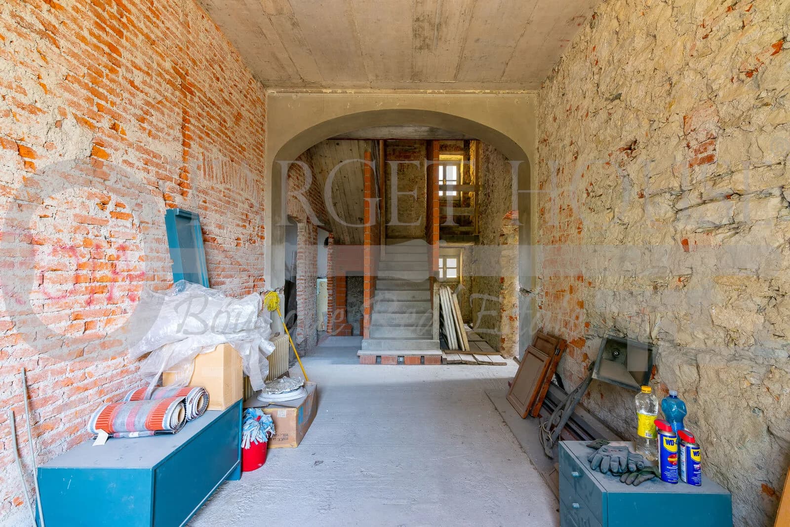Historic villa near Como renovation corridor with exposed brick walls, arched doorway, and concrete staircase under restoration