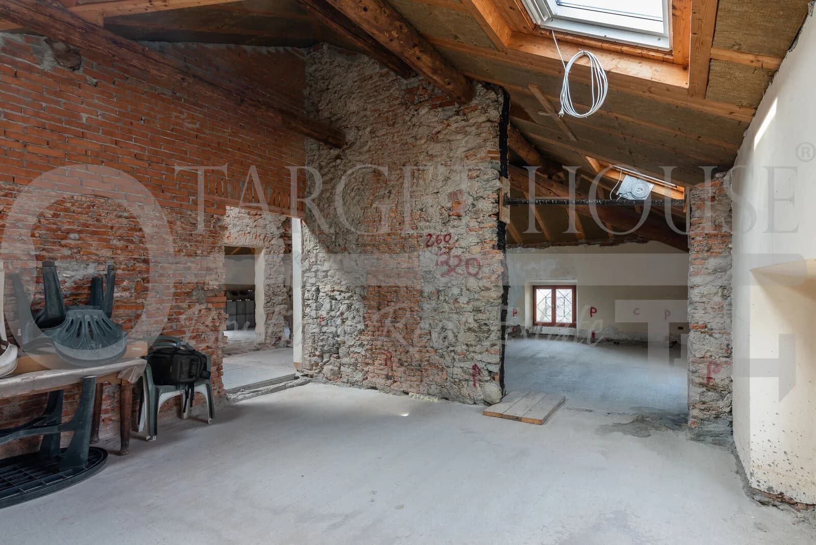 Unfinished attic with exposed brick walls, wooden roof beams, skylight, and concrete floors in historic villa near Como