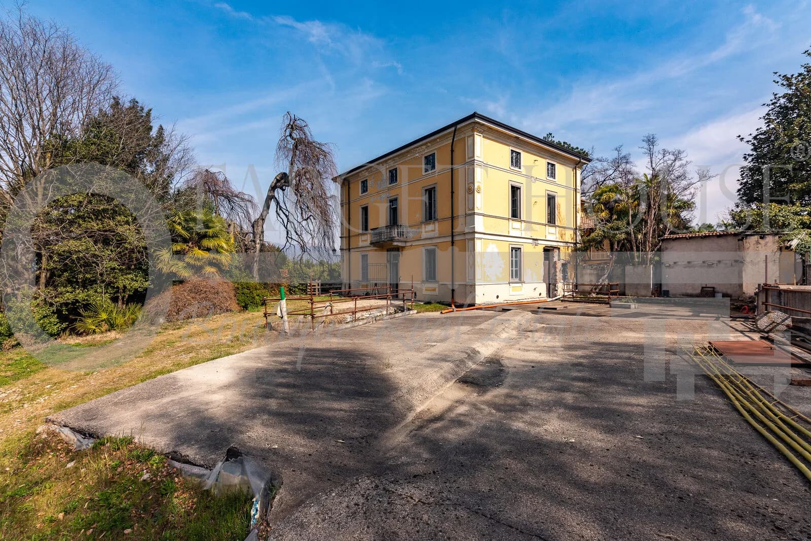 Historic villa near Como with yellow facade, large paved courtyard, lush garden, and mature trees under blue sky